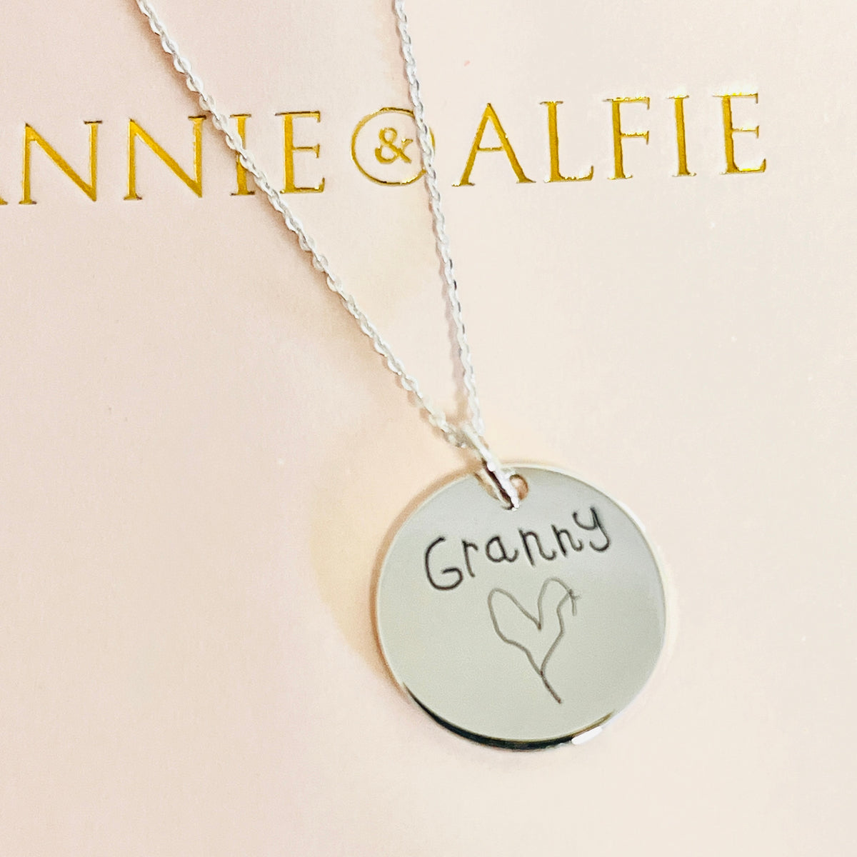 A silver round disc pendant necklace engraved with the handwriting 'Granny' and a heart, hanging from a silver chain against a pale pink background with gold 'ANNIE & ALFIE' branding at the top.